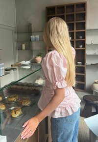 Femme aux longs cheveux blonds portant un chemisier rose et un jean, debout au comptoir d'une boulangerie avec des pâtisseries exposées.