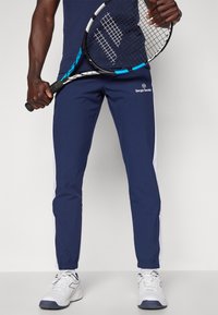 Marineblå atletiske bukser med hvide sidestriber, elastiske manchetter og et Sergio Tacchini-logo. Sammen med hvide atletiske sko.