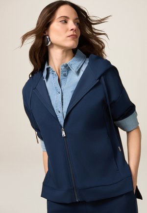 Marineblå zip-up hættetrøje lavet af et glat, strækbart stof, med hætte og korte ærmer over en lys blå denimskjorte.