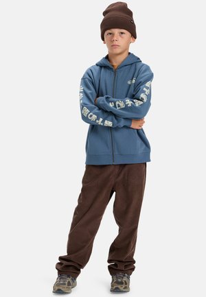 Blauwe rits-hoodie met logo-print op de mouwen, gecombineerd met bruine corduroy broek en een bruine beanie. Sneakers hebben een camouflagepatroon.