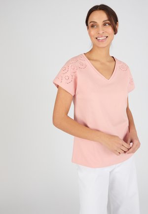 Vrouw lacht en draagt een roze V-hals top met korte mouwen met bloemen oogjespatronen op de schouders en witte broek.