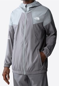 Kevyt, vetoketjullinen takki harmaana, jossa on kaksivärinen muotoilu. Ominaisuuksiin kuuluvat huppu, joustavat hihansuut ja sivutaskut. Logotähkät ovat läsnä.