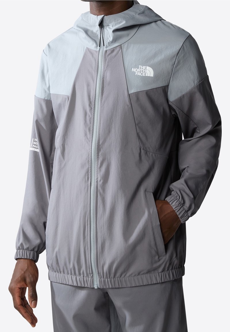 Kevyt, vetoketjullinen takki harmaana, jossa on kaksivärinen muotoilu. Ominaisuuksiin kuuluvat huppu, joustavat hihansuut ja sivutaskut. Logotähkät ovat läsnä.