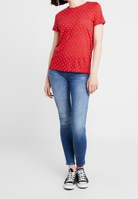 T-shirt rossa a maniche corte con pois bianchi e neri abbinata a jeans skinny blu chiaro e sneakers nere. Dettaglio del tessuto testurizzato visibile.