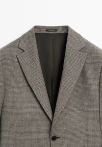 Grey plaid blazer with a notch lapel, single button closure, and a black interior lining. Features a textured fabric with a check pattern.