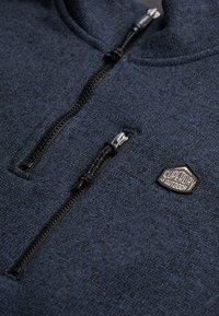 Dunkelblauer Fleece-Pullover mit hohem Kragen, verfügt über einen Reißverschluss vorne, schwarze Beschläge und eine strukturierte Oberfläche mit einem Markenpatch.