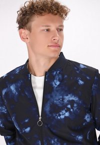 Marineblauwe bomberjack met een tie-dye patroon, voorzien van een ritssluiting aan de voorkant en een rond trekkoord, gemaakt van lichtgewicht stof.