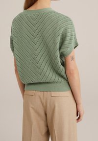 En person som bär en grön stickad kortärmad topp med ett zigzag-mönster och beige byxor, sedd bakifrån.