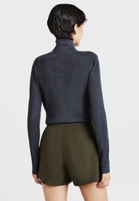 Persona con capelli corti e scuri, che indossa un maglione a collo alto grigio scuro a costine e pantaloni corti a vita alta verdi oliva, di spalle.