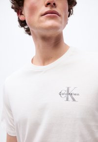 Νέος άνδρας που φοράει λευκό T-shirt Calvin Klein με λογότυπο CK στο στήθος, ουδέτερη έκφραση, κόπηκε κάτω από τα μάτια, ορατά σγουρά μαλλιά.