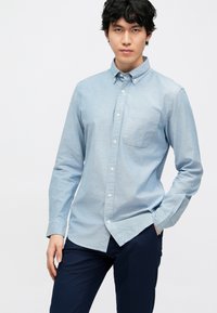 Camisa azul-claro de botão em tecido chambray, com um bolso no peito, mangas compridas e colarinho pontudo. Combinada com calças escuras.