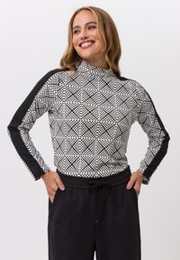 Glanzende vrouw met blond haar draagt een zwart-wit gestippeld shirt met lange mouwen en zwarte strepen, samen met zwarte broek met haar handen in de zakken.