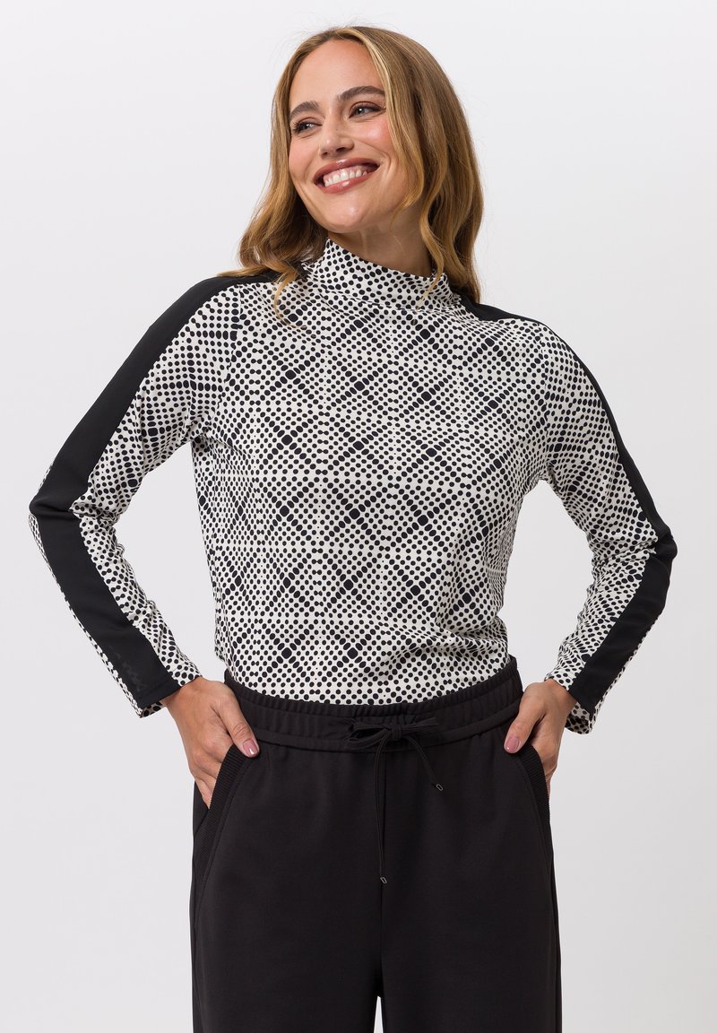 Glanzende vrouw met blond haar draagt een zwart-wit gestippeld shirt met lange mouwen en zwarte strepen, samen met zwarte broek met haar handen in de zakken.