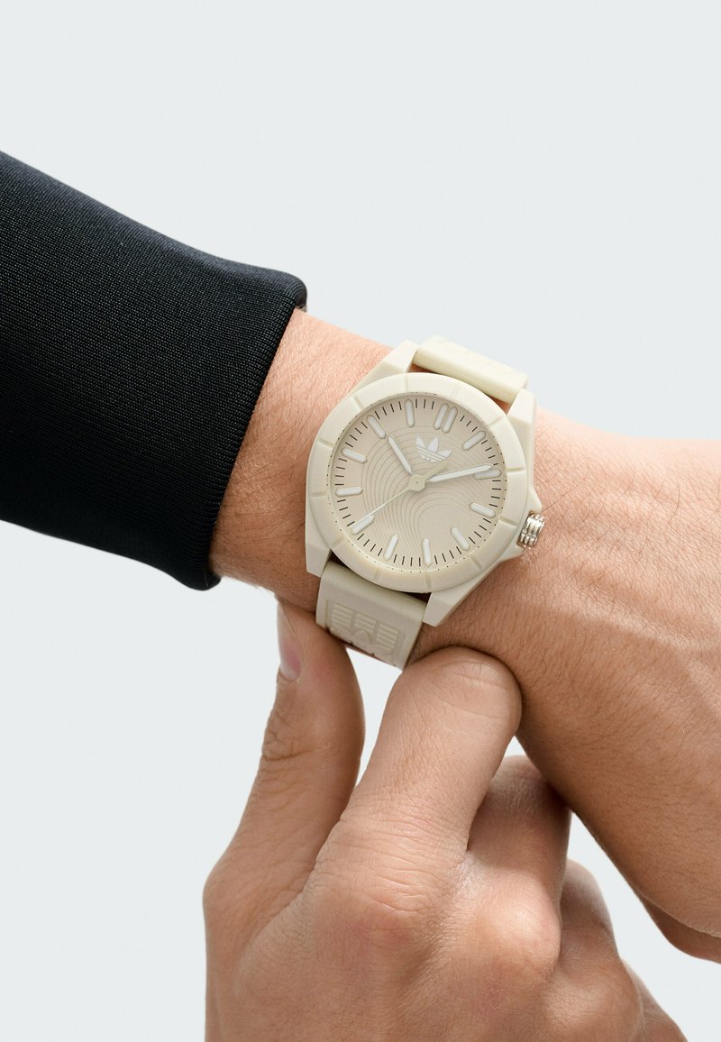 Montre beige clair avec cadran texturé, bracelet en caoutchouc à motifs et accents argentés. Portée au poignet avec une manche noire ajustée.