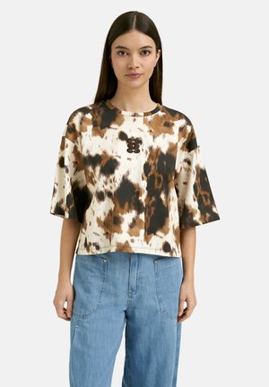 Frau mit langen dunklen Haaren trägt ein locker sitzendes kurzärmeliges Batikshirt in Braun, Schwarz und Weiß, kombiniert mit hellblauen Jeans.