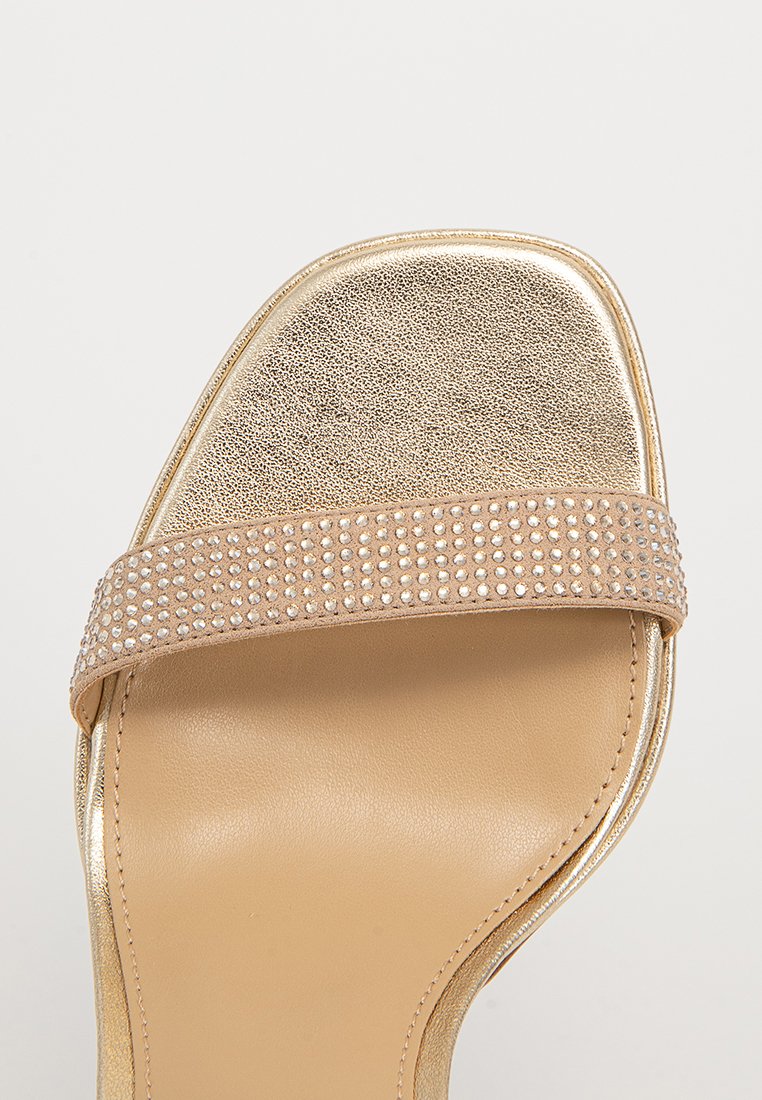 Sandale à glissière dorée avec une large sangle beige ornée de petits strass transparents. Semelle intérieure en cuir lisse, talon plat et bout rond.