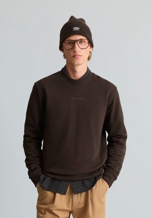 Giovane uomo con berretto marrone, grandi occhiali, felpa marrone scuro sopra una camicia a quadri nera e pantaloni beige, in piedi con le mani in tasca.