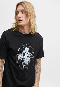 Ung person med langt blondt hår og tatoveringer iført en sort t-shirt med hvid blomstermønster og teksten "Aria Elegance Originals."