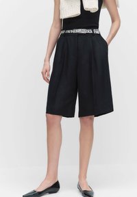 Mustat pleiattut shortsit, jotka on valmistettu kevyestä kankaasta, sivutaskuilla ja kuvioidulla vyöllä. Yhdistetty mustiin kärjellisiin ballerinoihin.