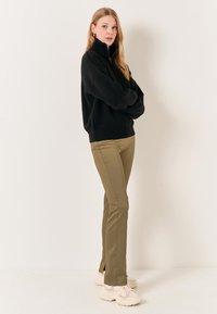 Schwarzer Strickpullover mit hohem Kragen, kombiniert mit olivgrünen Slim-Fit-Hosen und hellen Sneakers, vor einem einfarbigen Hintergrund stehend.