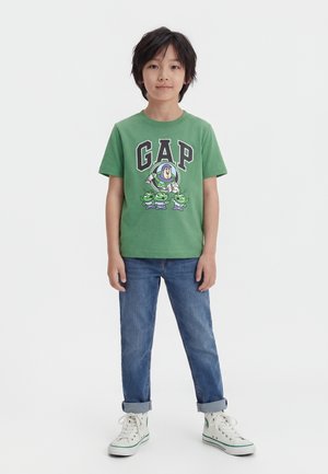 Παιδί που στέκεται, φοράει πράσινο μπλουζάκι με εκτύπωση του Buzz Lightyear και τρεις πράσινους εξωγήινους, μπλε τζιν και λευκά ψηλά αθλητικά παπούτσια.