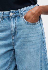 Άτομο που φοράει ανοιχτό μπλε τζιν παντελόνι με το δεξί χέρι μερικώς μέσα στην εμπρόσθια τσέπη, σε απλό φόντο.