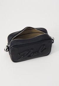 Sac en cuir noir avec une finition texturée, de forme rectangulaire et logo en relief bien en vue. Il est doté d'une fermeture éclair argentée et d'une doublure intérieure.