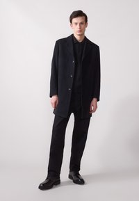 Manteau en laine noir avec revers, fermeture à boutons et poches latérales, associé à un polo noir et un pantalon noir. Texture lisse.