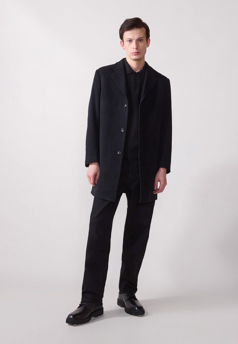 Manteau en laine noir avec revers, fermeture à boutons et poches latérales, associé à un polo noir et un pantalon noir. Texture lisse.