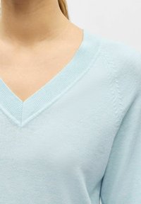 Gros plan d'une personne portant un pull bleu clair à col en V avec des détails subtils en motif tricoté sur l'épaule.