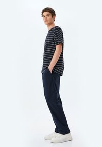 Stribet navy t-shirt med korte ærmer, parret med navy bukser og hvide sneakers, der står foran en enkel lys baggrund.