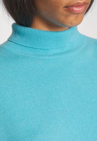 Pull à col roulé bleu canard en tissu côtelé. Vue en gros plan mettant en avant la texture et le design du col ainsi que le motif du tricot.