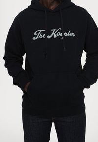 Muž v černé mikině s kapucí, na hrudi s vyšitým nápisem "The Kooples", má ruce v přední kapse a je oblečen v tmavých džínách.