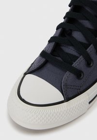 Platneni tenisice tamno sive boje s crnim vezicama, bijelim gumastim nosom i teksturiranim potplatom. Imaju kontrastnu obojenu srednju podlogu i rupice za vezice.