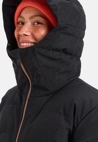 Zwarte pufferjack met een hoge kraag en capuchon, met een koperen rits. Draag het boven een rode geribde beanie. Geweven stof met een doorgestikte afwerking.