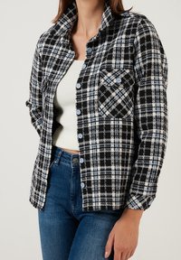 Camicia a quadri bianca e nera con bottoni blu, dotata di colletto e taschino frontale; realizzata in un tessuto testurizzato. Indossata sopra una maglietta bianca.