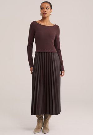 Femme portant un haut marron foncé à manches longues, une jupe maxi plissée assortie, des bottes en daim beige et des boucles d'oreilles créoles en or, debout devant un fond neutre.
