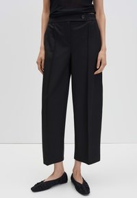 Vrouw in zwarte wijde cropped broek met een knoopsluiting en zwarte gerimpelde ballerina's voor een effen achtergrond.