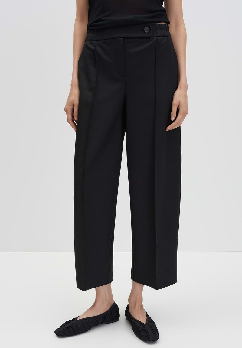 Vrouw in zwarte wijde cropped broek met een knoopsluiting en zwarte gerimpelde ballerina's voor een effen achtergrond.