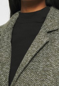 Blazer verde texturizado con solapa a medida, combinado con un cuello de tortuga negro ajustado y acanalado. Primer plano, mostrando detalles y patrones de la tela.