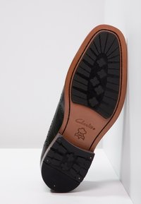 Semelle de chaussure en cuir noir avec des détails de brogue complexes ; semelle intermédiaire marron ; semelle extérieure en caoutchouc noir avec un motif de grip texturé.