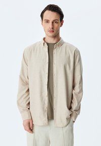 Camisa de botones color beige claro, hecha de tela suave, con collar y mangas largas, combinada con una camiseta verde. Pantalones color crema.