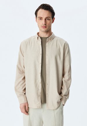 Camisa de botones color beige claro, hecha de tela suave, con collar y mangas largas, combinada con una camiseta verde. Pantalones color crema.