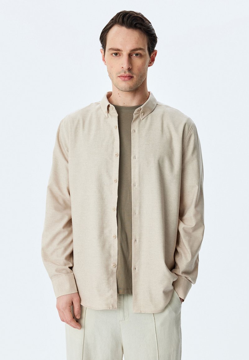 Camisa de botones color beige claro, hecha de tela suave, con collar y mangas largas, combinada con una camiseta verde. Pantalones color crema.
