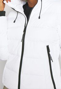 Hvid dunjakke med lynlås foran, sorte detaljer, hætte og to lynlåslommer. Stoffet virker glat med en polstret tekstur.