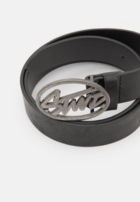 Zwarte leren riem met een ronde, zilverkleurige gesp met een cursieve logoontwerp. De riem heeft een gladde textuur en strakke lijnen.