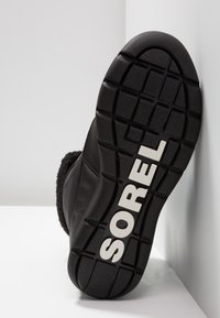 Black boot sole with rubber tread and white "SOREL" branding. Features a textured pattern for grip and a soft, dark fur-like upper detail.