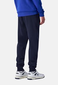 Marineblauwe sweatpants van zacht materiaal met taps toelopende pijpen en ribgebreide boorden, gecombineerd met witte sportschoenen met zwarte en grijze accenten.