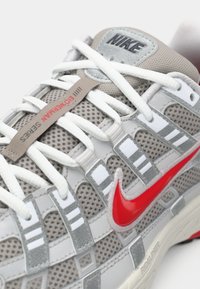 Nike-Sneaker aus grauem Mesh mit rotem Swoosh. Verfügt über weiße Schnürsenkel, graue Überzüge und ein strukturiertes Muster an den Seiten. Hardware-Tag sichtbar.