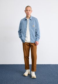 Chemise boutonnée bleu clair avec poche poitrine, associée à un pantalon marron clair et des baskets de couleur claire. Couleurs unies, coupe décontractée.